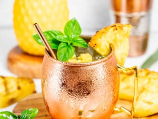 A frosty copper mug filled with a tropical drink, garnished with a pineapple slice and fresh basil. In the background, a pineapple, a cocktail shaker, and lime wedges are visible on a wooden board.