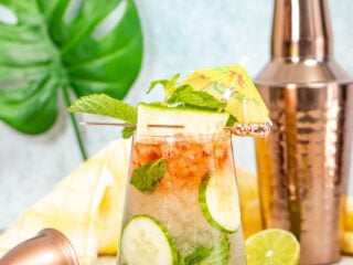 A refreshing cocktail with lime, cucumber slices, and mint, garnished with a pineapple slice and paper umbrella, sits on a wooden coaster. Behind it are a copper shaker, jigger, a cut lime, and a green leaf decor.