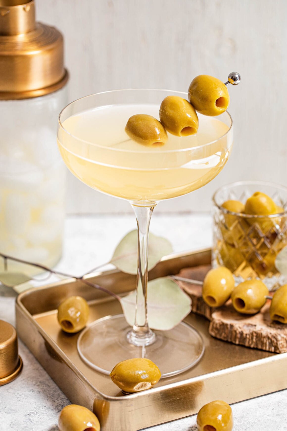 A coupe glass brimming with a pale yellow, elegantly garnished with green olives on a skewer, rests on a gold tray surrounded by more olives.